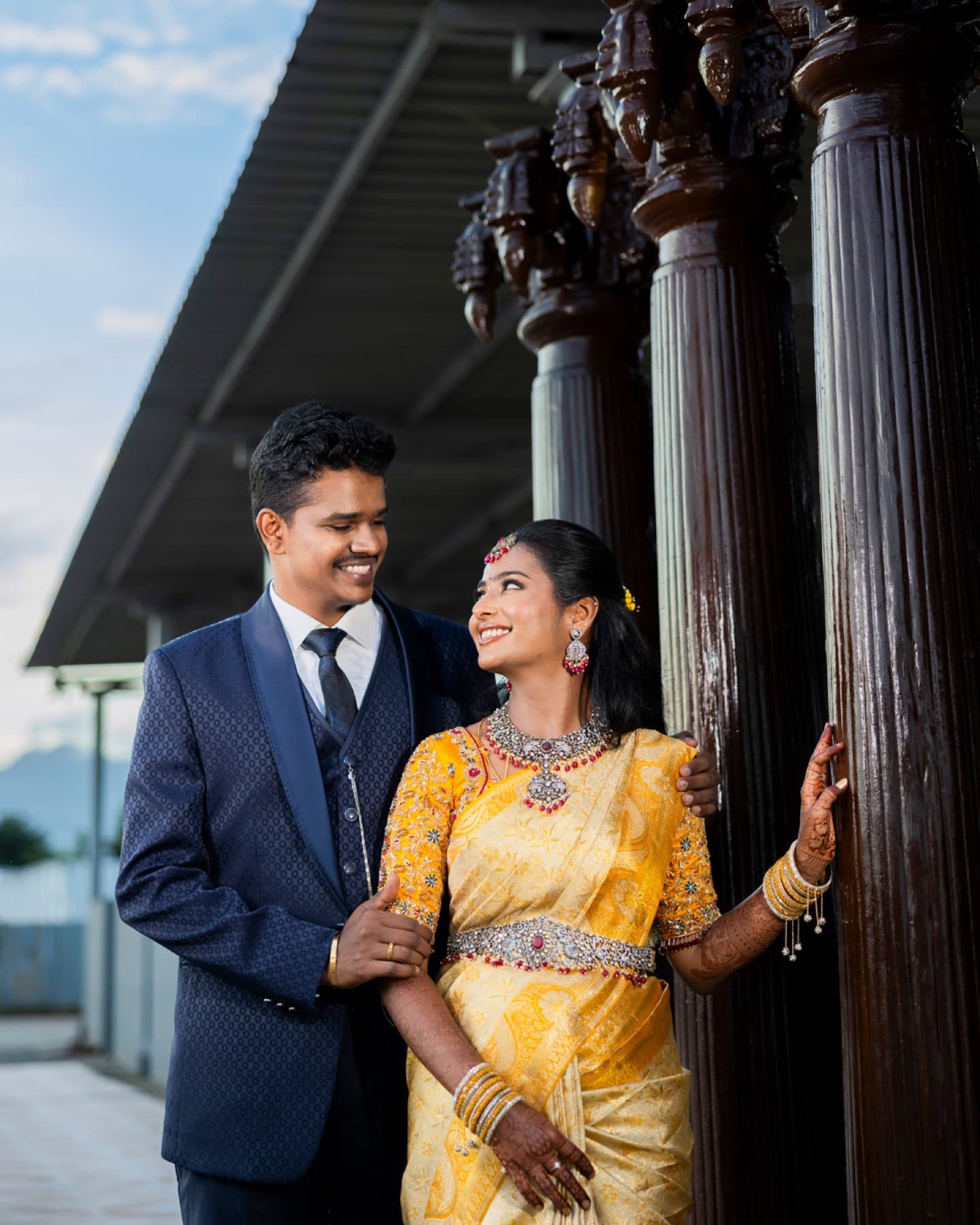 Couple in a suit and yellow silk saree beside wooden pillars